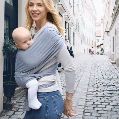 Jeune-maman-se-promenant-dans-une-rue-piétonne-portant-dans-son-écharpe-de-portage-bébé-gris-clair-son-bébé-apaisé.