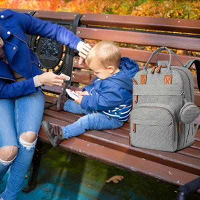 Bébé-jouant-avec-sa-maman-sur-un-banc-dans-un-parc-à-côté-duquel-se-trouve-son-sac-à-dos-à-langer-gris-sur-lequel-est-accroché-une-mini-sacoche-grise-amovible