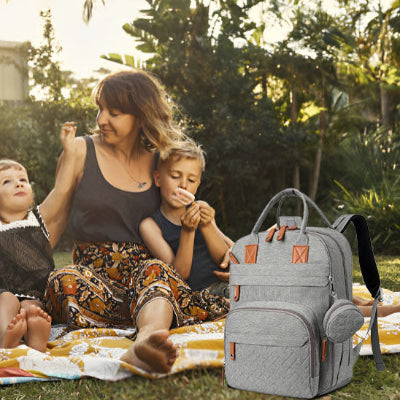 Maman-en-pique-nique-avec-ses-enfants-dans-un-jardin-à-côté-desquels-est-posé-son-sac-à-dos-à-langer-gris-à-fermetures-éclair