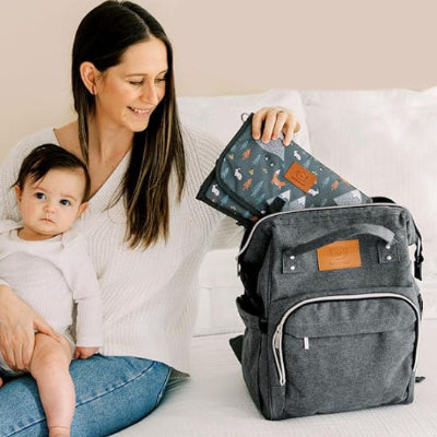 Bébé-assis-sur-les-genoux-de-sa-maman-assise-sur-son-canapé-sur-lequel-se-trouve-son-sac-à-langer-gris-à-sangles-ajustables-et-clipsablles