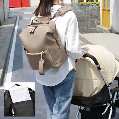 Jeune-maman-portant-une-chemise-blanche-sur-laquelle-se-trouve-son-sac-à-langer-de-couleur-caramel-contenant-une-poche-accueillant-son-téléphone-porté-façon-sac-à-dos-en-train-de-marcher-derrière-sa-poussette-dans-une-rue-étroite-d-un-centre-ville-lors-d-une-journée-d-été-ensoleillée