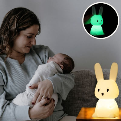 Maman-installée-dans-son-canapé-tenant-dans-ses-bras-son-bébé-endormi-à-côté-duquel-est-posé-sa-veilleuse-bébé-en-forme-de-lapin-rassurante-et-reconfortante